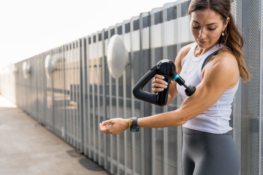 woman in workout clothes using theragun on her arm 